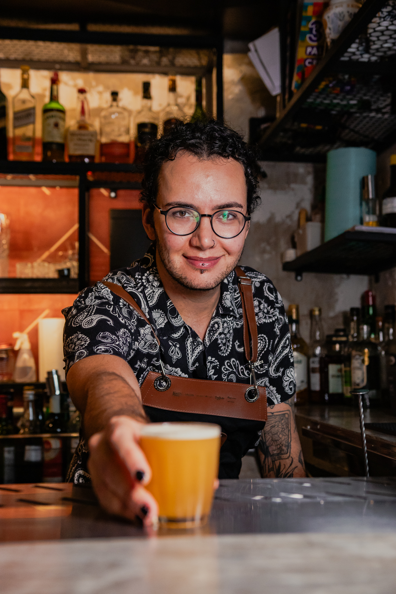 O bartender, Aleksei Borges, empurra com um sorriso um copo pequeno no modelo "barriquinha" com o coquetel Macunaíma em direção à câmera.