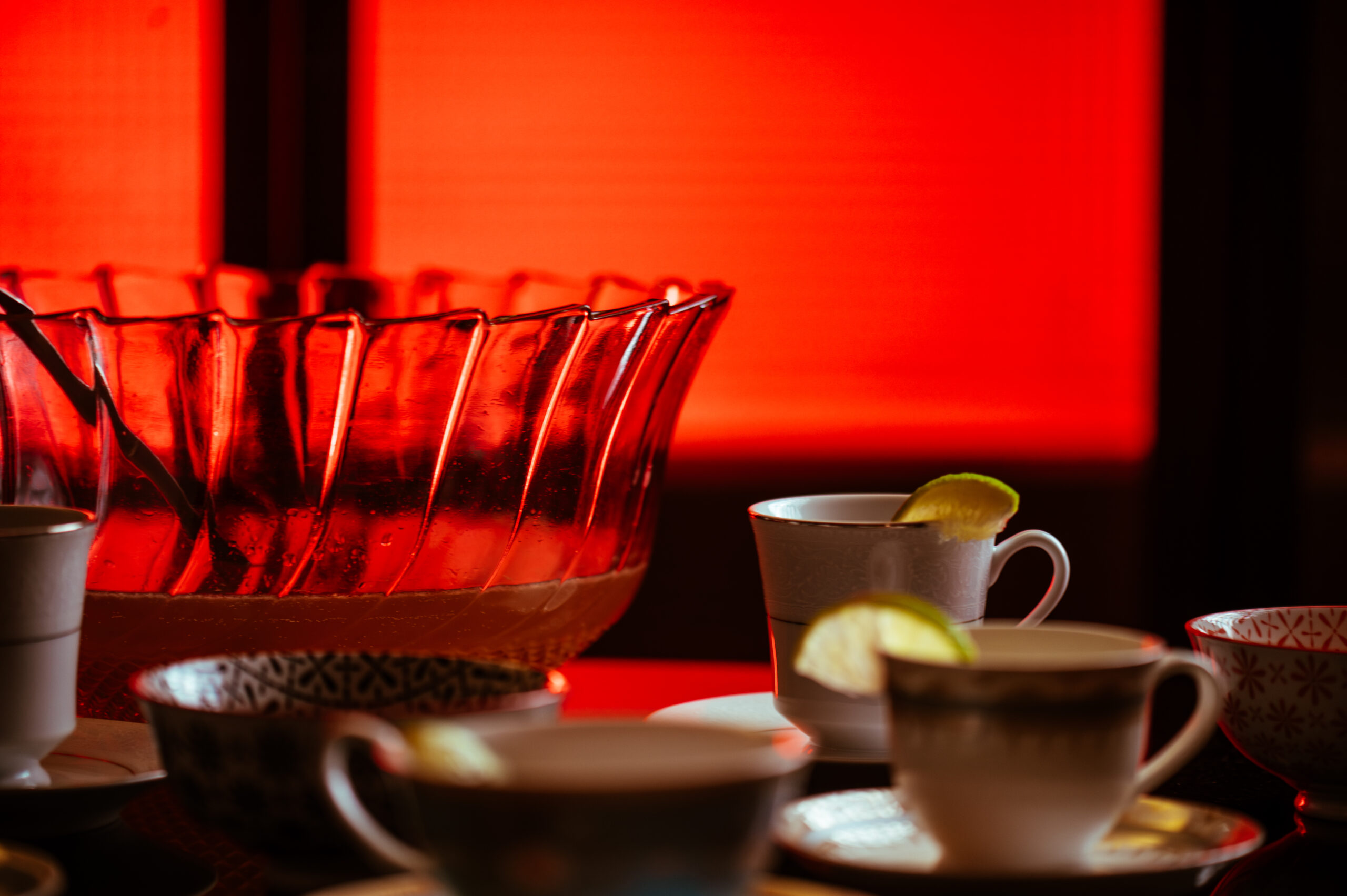 À frente da imagem, vê-se diversas xícaras de chá de porcelana, em diferentes modelos. Um pouco atrás, à esquerda, um grande bowl de punch de vidro lapidado, cheio de punch e com uma concha de servir prateada dentro. Toda a iluminação da foto é em tons de vermelho.